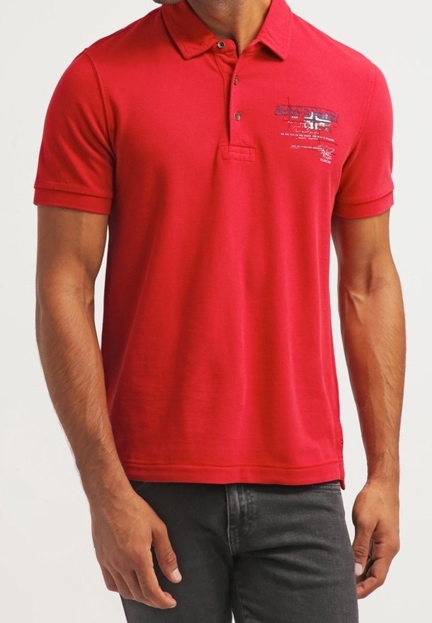 Homme portant un polo rouge à manches courtes avec un petit motif et un texte sur le côté gauche de la poitrine, assorti à un jean foncé.