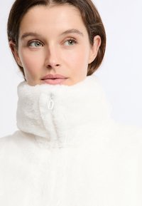 Bia�ła, pluszowa kurtka z wysokim kołnierzem i zamkiem błyskawicznym z przodu. Miękka faktura, minimalistyczny design oraz brak widocznych wzorów czy detali hardware.