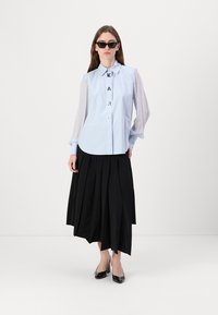 Camicia azzurra chiara con colletto strutturato e maniche trasparenti; abbinata a una gonna nera plissettata e tacchi neri lucidi.