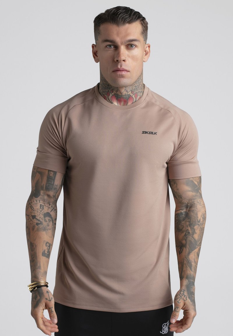 Homme avec des tatouages, y compris un tatouage au cou, portant une chemise beige à manches courtes SIKSILK, debout devant un fond gris uni.