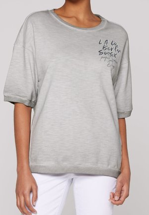 Kobieta ubrana w jasnoszarą koszulkę z krótkim rękawem z czarnym napisem w górnej lewej części oraz białe spodnie, pokazana od szyi do bioder.