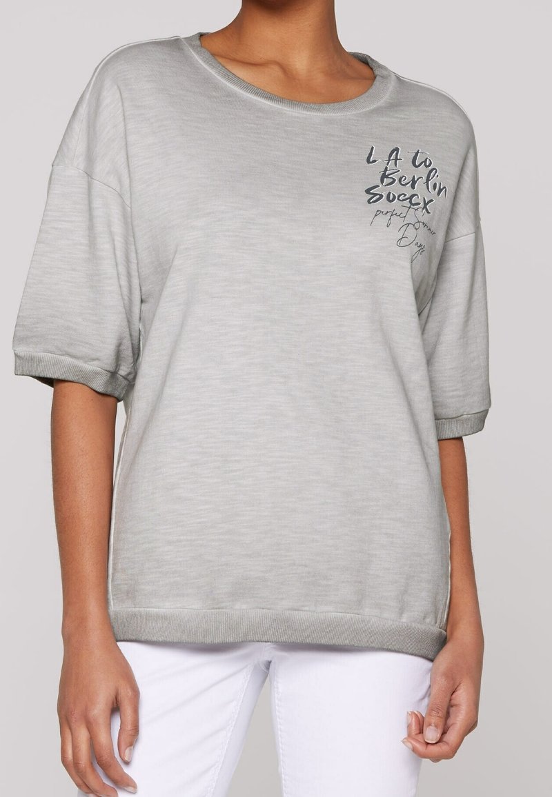 Femme portant une chemise à manches courtes gris clair avec un texte noir sur le côté supérieur gauche et un pantalon blanc, montrée du cou aux hanches.