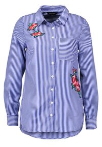 Chemise à manches longues rayée verticalement bleu et blanc avec poche avant et broderie florale rose sur la poitrine et le bas côté.