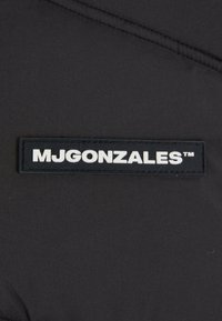 Zwarte stof met een verhoogd rechthoekig etiket met "MJGONZALES™" in witte letters, dat een gladde textuur en minimalistisch ontwerp benadrukt.