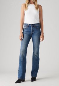 Jeans acampanados en denim azul medio con lavado desgastado, talle alto y detalles sutiles de costura, combinados con una camiseta de tirantes blanca básica.