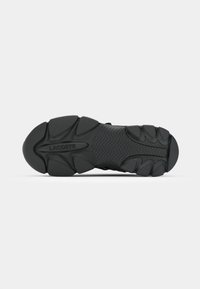 Semelle de chaussure de sport noire en caoutchouc texturé, présentant un motif en zigzag et des détails de bande de roulement prononcés. Nom de la marque "LACOSTE" embossé.