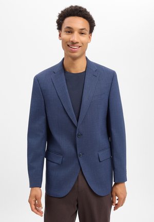 Giovane uomo sorridente, indossa un blazer blu a due bottoni sopra una camicia scura e pantaloni marroni, in piedi davanti a uno sfondo chiaro e semplice.