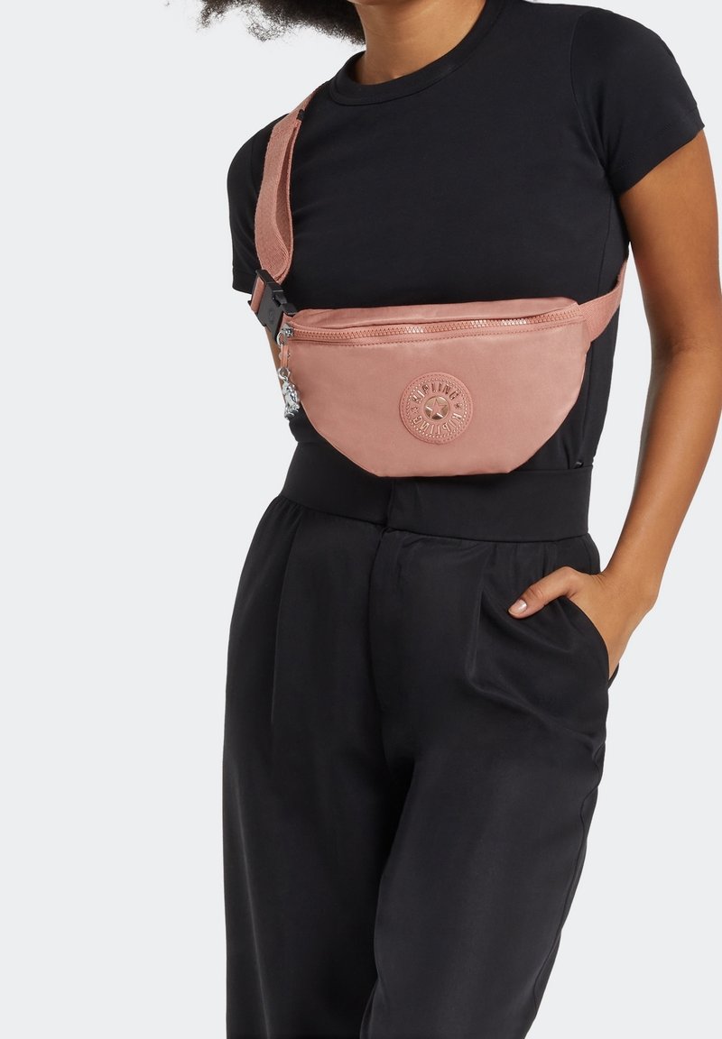 Personne portant un haut noir à manches courtes et un pantalon avec un sac banane rose à bandoulière, orné d'un patch logo rond et d'une fermeture éclair.