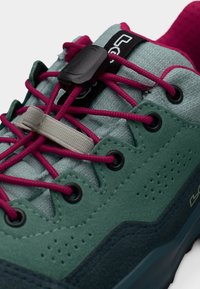 Close-up van een blauwgroene en donkerblauwe sneaker met roze veters die vastzitten met een zwart schuifslot en een beige veterhouder op een lichtgrijze tong.