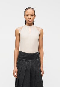 Γυναίκα με πλεκτά μαλλιά, που φοράει μπεζ αμάνικο τοπ με δαντέλα στους ώμους και σκουρόχρωμη πλισέ φούστα ψηλόμεση με πλευρικά λουράκια.