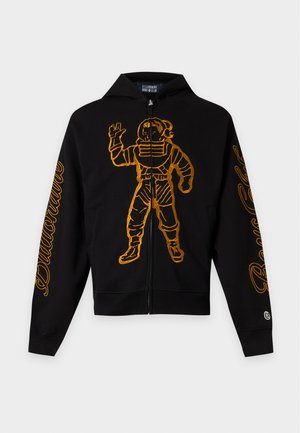 Mladý muž v čiernej mikine na zips s žltou grafikou astronauta, voľné svetlomodré rifle, sivé tričko a čierno-biele tenisky stojí s rukami v vreckách.
