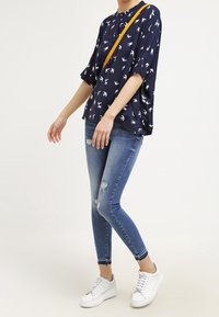 Blouse bleu marine avec imprimé d'oiseaux blancs, coupe ample, manches courtes. Portée avec un jean bleu déchiré et des baskets blanches, avec une bandoulière jaune.