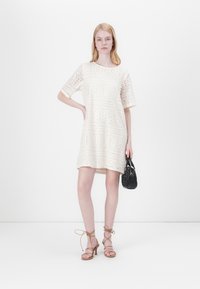 Abito in pizzo all'uncinetto di colore bianco sporco, dal fit morbido, con maniche corte e pattern floreali. Abbinato a sandali nudi con cinturino e una piccola borsa nera.
