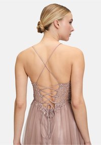 Długa sukienka w odcieniu blush z misternym haftem kwiatowym na plecach, z detalami w postaci związanych ramiączek i zwiewną, przezroczystą fakturą.