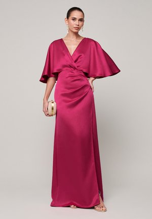 Vrouw die een lange magenta satijnen jurk draagt met brede capemouwen, een kleine gouden clutch vasthoudt en voor een effen achtergrond staat.