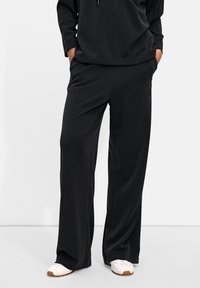 Black, loose-fitting trousers with a smooth texture and elastic waistband, featuring side pockets and a subtle logo detail.