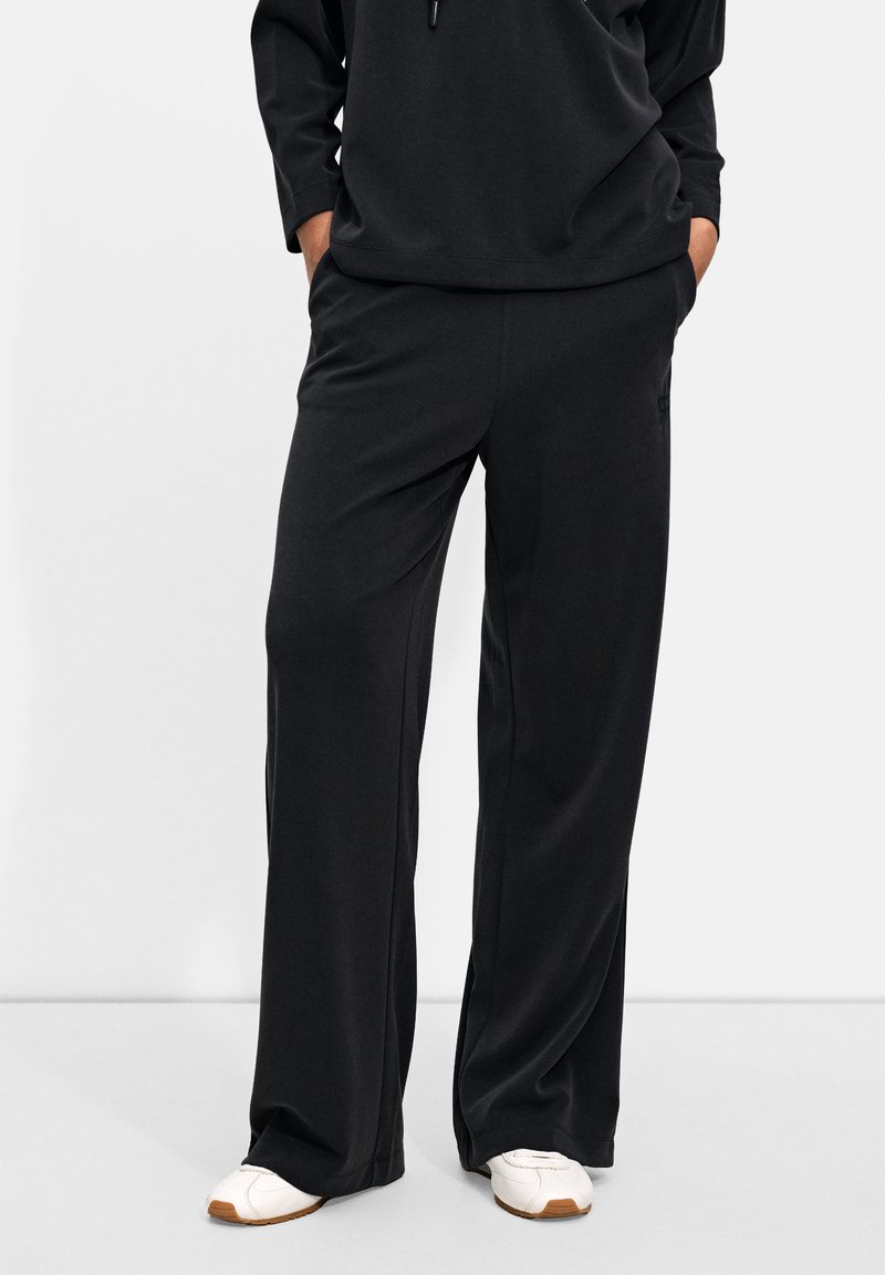 Black, loose-fitting trousers with a smooth texture and elastic waistband, featuring side pockets and a subtle logo detail.