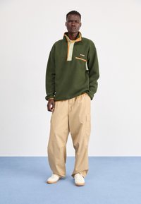 Grøn fleecepullover med orange krave og knapper, der har en frontlomme. Kombinationen er med cremefarvede cargo-bukser og hvide sneakers.