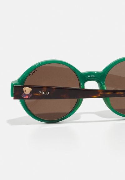 Lunettes de soleil rondes avec une monture verte pétillante et des verres marron. Les branches arborent un motif écaille de tortue et un logo d'ours avec un détail de polo.