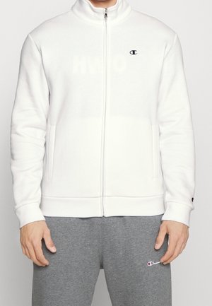 Person iført en hvid lynlåstrøje med et lille logo på brystet og grå joggingbukser med et logo nær lommen.