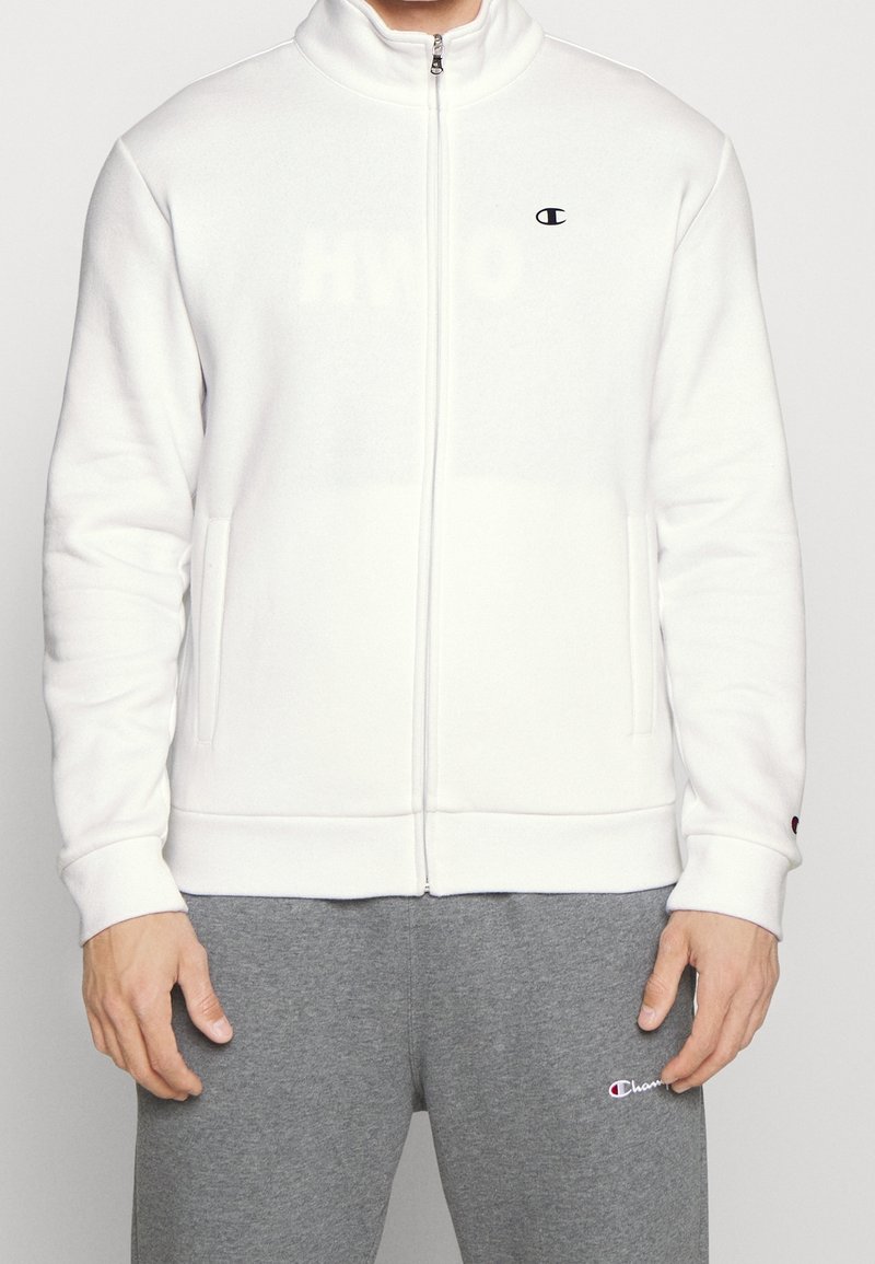 Personne portant une veste blanche zippée avec un petit logo sur la poitrine et un pantalon de survêtement gris avec un logo près de la poche.