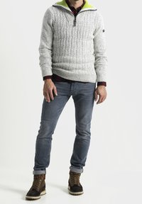 Maglione grigio a trecce con mezzo zip e colletto verde brillante, abbinato a jeans blu e stivaletti marroni con lacci.