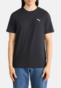 Schwarzes Baumwoll-T-Shirt mit kurzen Ärmeln und rundem Ausschnitt, mit einem kleinen weißen Puma-Logo auf der linken Brust. Kombiniert mit blauen Jeans.