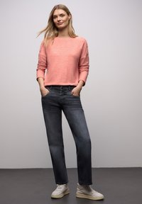 Jasnoróżowy dzianinowy sweter z podwiniętymi rękawami, w połączeniu z ciemnoszarymi prostymi jeansami i białymi sneakersami. Modelka stoi z rękami w kieszeniach.