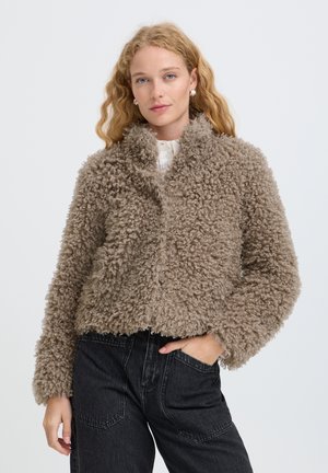Femme aux longs cheveux blonds bouclés portant des boucles d'oreilles argentées, une veste texturée bouclée taupe, une chemise blanche et un jean noir sur fond blanc.