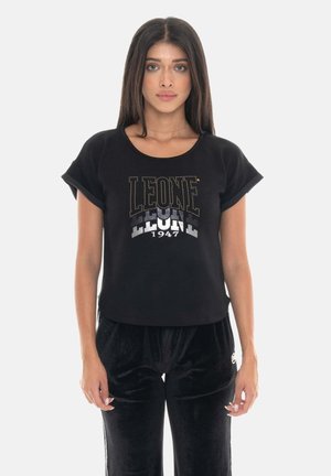 Donna che indossa una maglietta nera con la scritta "LEONE 1947" e pantaloni in velluto neri, in piedi davanti a uno sfondo bianco semplice.