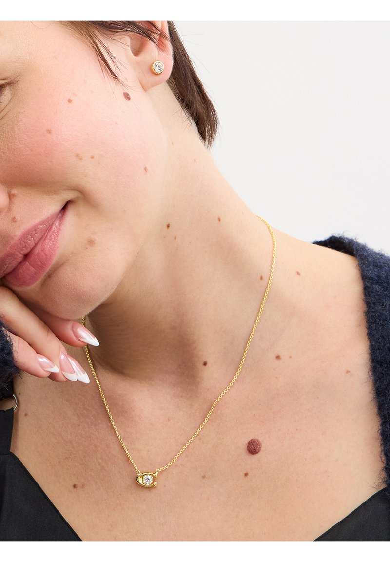 Mujer inclinando la cabeza con un collar dorado que tiene un pequeño colgante y pendientes de botón a juego, vistiendo una blusa negra y un cárdigan oscuro.