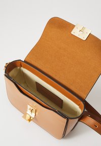 Sac bandoulière en cuir tan avec un fermoir en métal doré. L'intérieur est en daim tan avec une doublure beige lisse et une étiquette de marque.