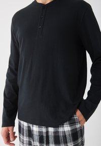 Chemise henley noire à manches longues avec trois boutons au cou, associée à un pantalon de pyjama à carreaux gris, présentant une texture lisse.