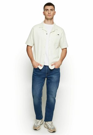 Ung mand står med hænderne i lommerne iført kortærmet hvid skjorte, hvid t-shirt, blå jeans og beige-hvide sneakers.