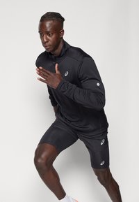 Svart atletisk långärmad topp med hög krage och logotyp, tillsammans med svarta kompressionsshorts. Slät tyg, åtsittande design.