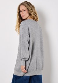 Kobieta o długich kręconych blond włosach, ubrana w szary, oversize'owy sweter z cekinami z przodu oraz szerokie niebieskie dżinsy.