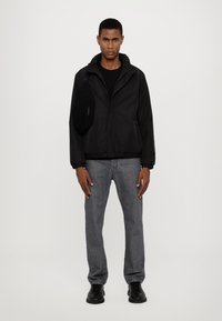 Giacca nera con zip e cappuccio, dotata di tasca laterale, abbinata a jeans grigi e stivali neri. Texture liscia e design casual.