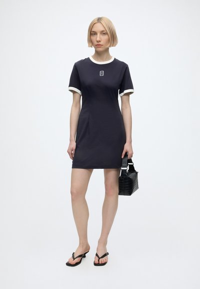Jeune femme aux cheveux blonds courts portant une robe noire à manches courtes avec un bord blanc, tenant un sac à main noir et portant des sandales à talons noires.