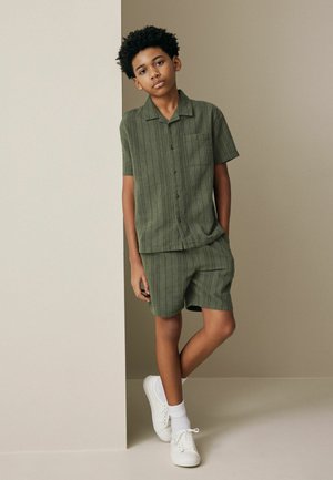 Boy with short curly hair stands against neutral wall, wearing green short-sleeve button-up shirt, matching shorts, white socks, and white sneakers.