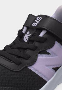 Κοντινό πλάνο μαύρου και άσπρου αθλητικού παπουτσιού με μαύρα κορδόνια, λουράκι Velcro και λεπτομέρειες λογότυπου στη γλώσσα και στην πλευρική επιφάνεια.