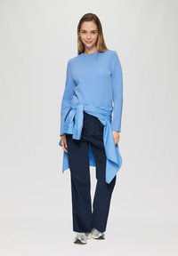 Lichtblauwe langemouwen top met ronde hals, gecombineerd met donkerblauwe wijdvallende broek. Een blauwe overhemd die om de taille is geknoopt. Zilveren sportschoenen.