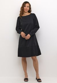 Zwarte jurk met lange mouwen, ronde halslijn en geplooide rok. Gemaakt van een gladde stof met een comfortabele pasvorm en elastische manchetten.
