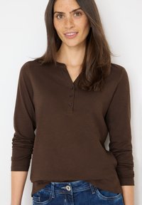Braunes langärmliges Henley-Shirt mit einer Drei-Knopf-Blende, abgerundetem Saum und weicher, glatter Textur, kombiniert mit blauen Jeans.