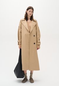 Beige trench coat with large lapels and double-breasted buttons. Model holds a black tote bag. Brown sweater and beige footwear complete the look.