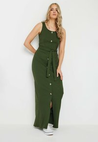 Robe longue olive verte avec une texture côtelée, dot�ée d'un col rond, de boutons dorés, d'une ceinture à nouer et d'une fente sur le côté. Portée avec des baskets blanches.