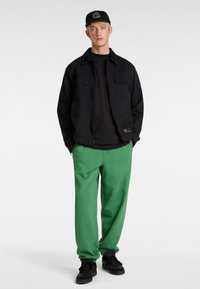 Schwarze Jacke über einem schwarzen Hemd, kombiniert mit grünen Hosen und schwarzen Sneakers. Die Jacke verfügt über zwei Fronttaschen und einen Reißverschluss.