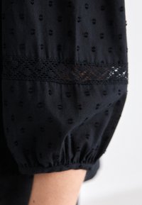 Manche en tissu noir avec des points texturés, bordure en dentelle et poignet élastique reposant sur un avant-bras sur un fond uni.