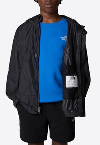 Svart lättviktig jacka med huva, med en meshficka. Under jackan en blå sweatshirt med logotyp, tillsammans med svarta shorts.