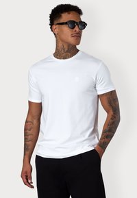 T-shirt blanc à col rond avec des manches courtes, fabriqué en coton, avec un petit logo brodé sur la poitrine, texture lisse, porté avec des lunettes de soleil noires.