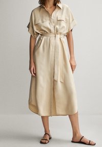 Beige satiininen paitamekko, jossa on lyhyet hihat, rintataskut ja siihen sopiva vyö. Mekossa on rento istuvuus ja polvenpituinen malli.